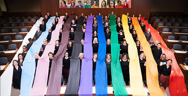 A large group of people holding rainbow colored banners form a pride flag.