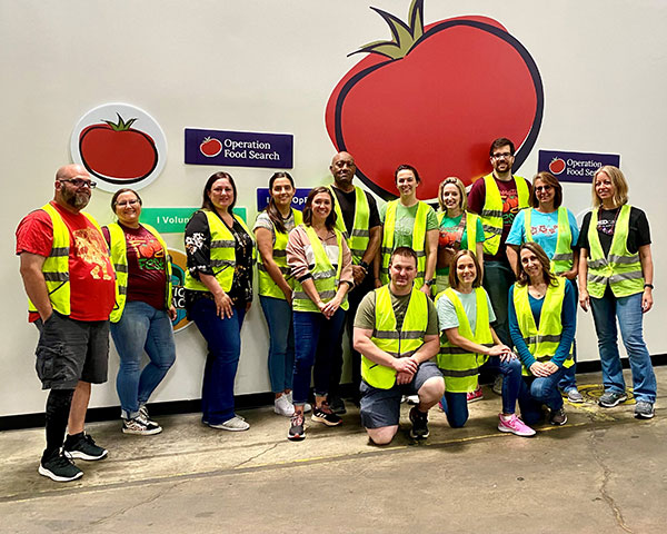 Several people pose in front of a wall with the Operation Food Search logo, which is a large tomato.